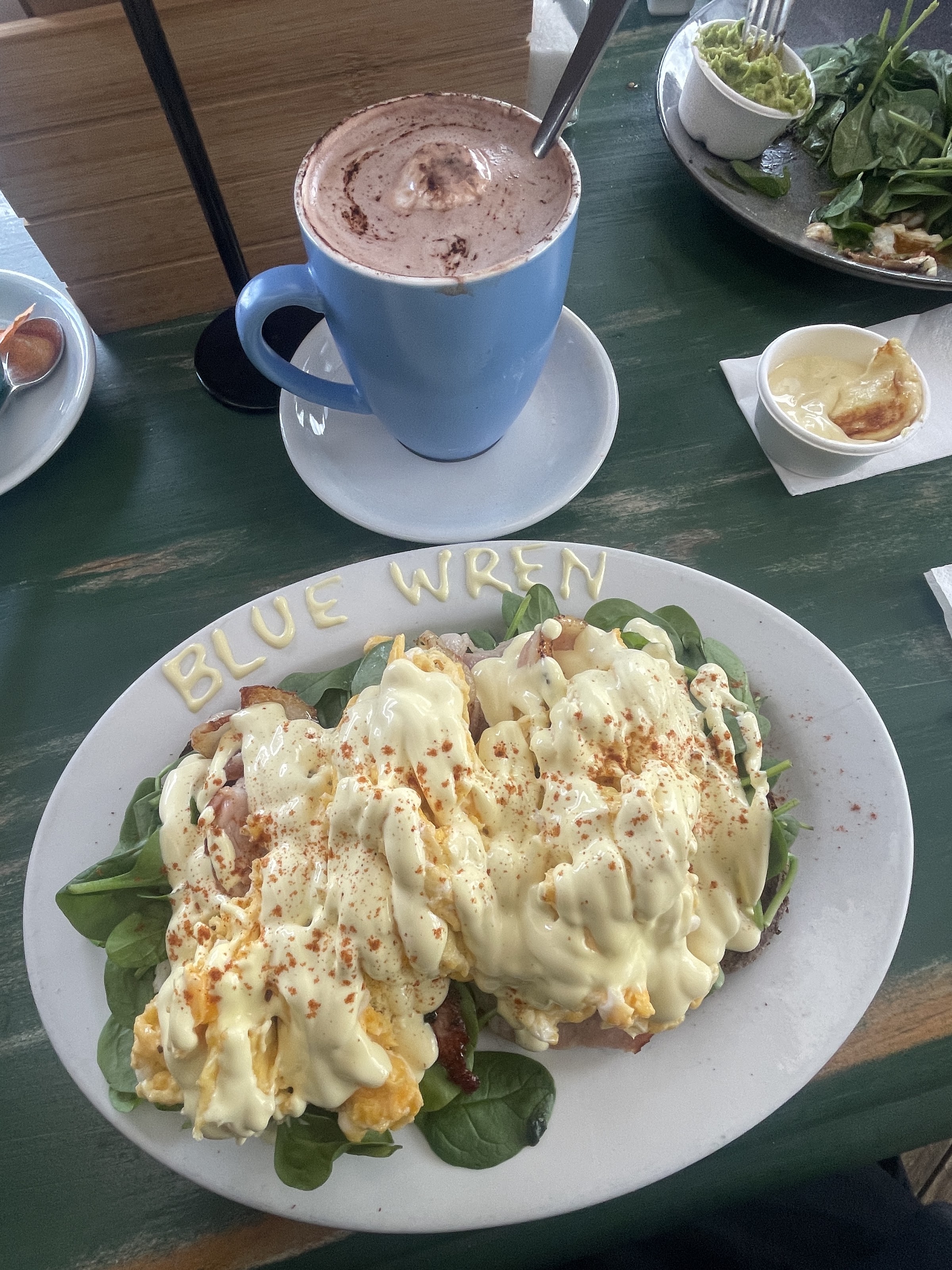 Eggs hollandaise on spinach with Blue Wren piped on the plate rim, beside a hot chocolate